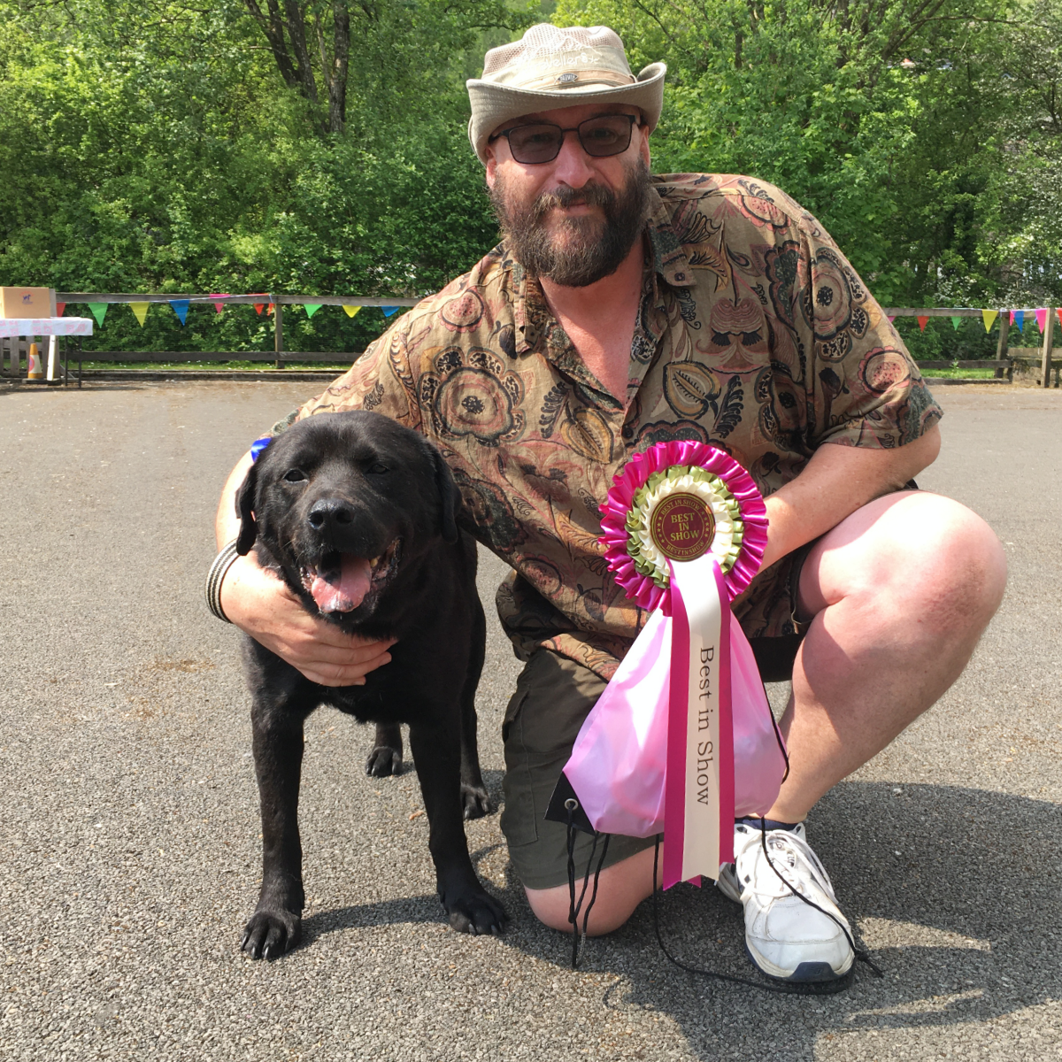 Bearded man wearing a hat cuddling a black labrador dog and holding a pink and white rosette